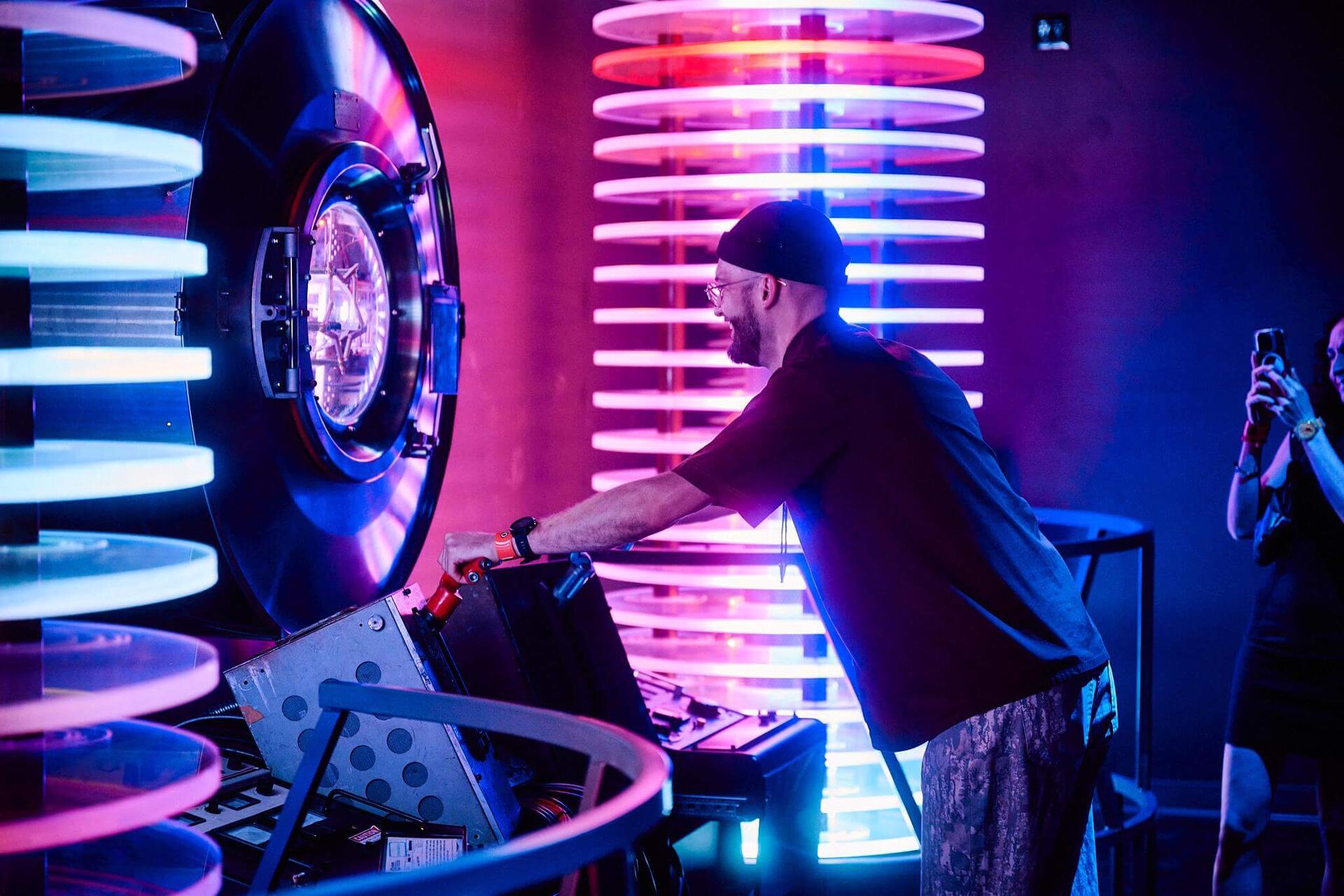 A performer working a console between towering illuminated ring sculptures glowing pink and blue