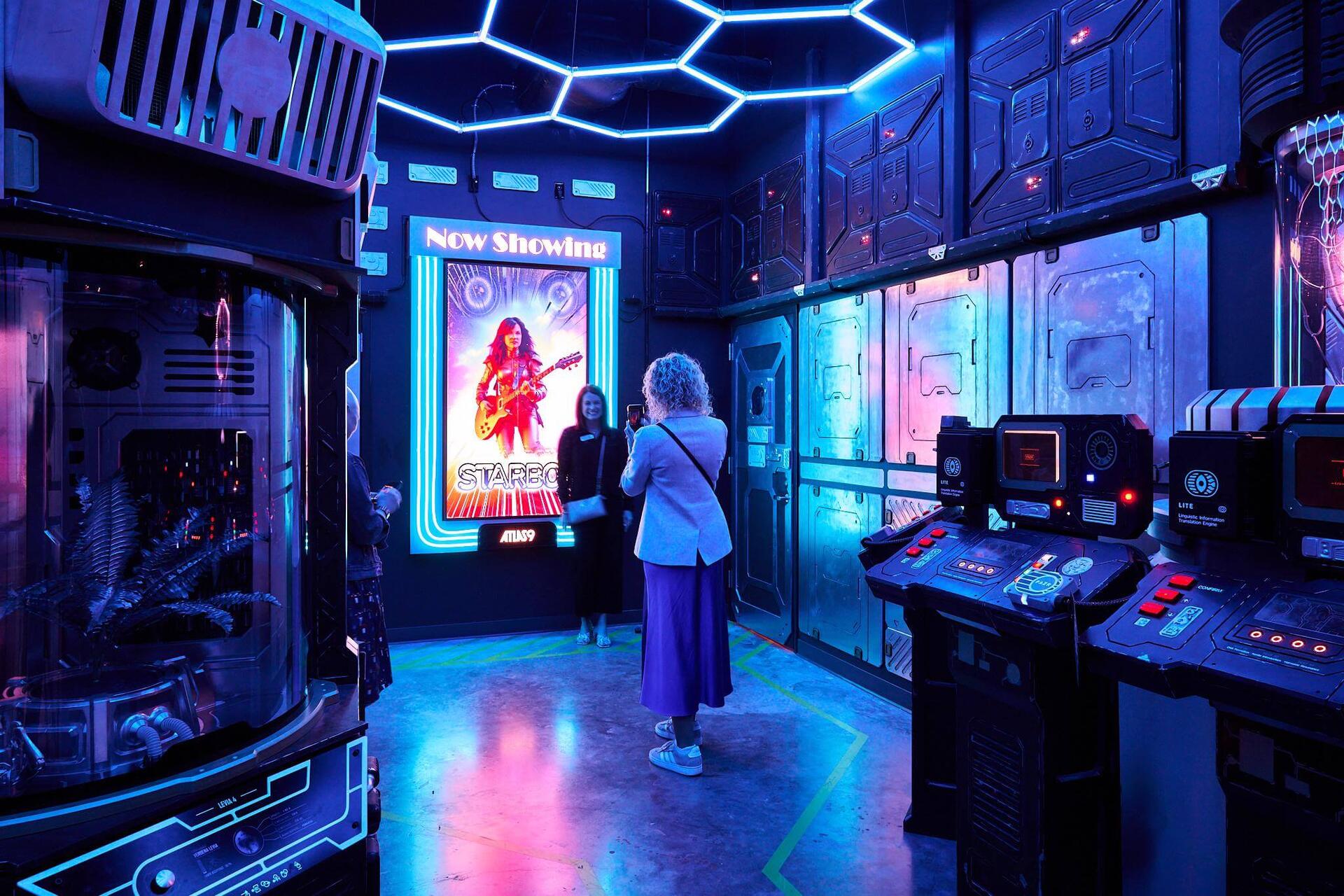Guests exploring a sci-fi themed room with neon hexagon ceiling, control panels, and a backlit movie poster